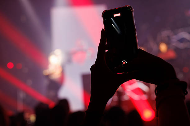Silhouette of a hand holding a phone while recording a live music performance. Red lights and bokeh create dynamic mood.