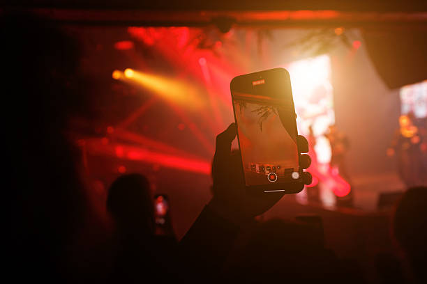 A person records a concert using a smartphone, capturing the stage and lighting effects. Red and orange tones create a dramatic concert mood.