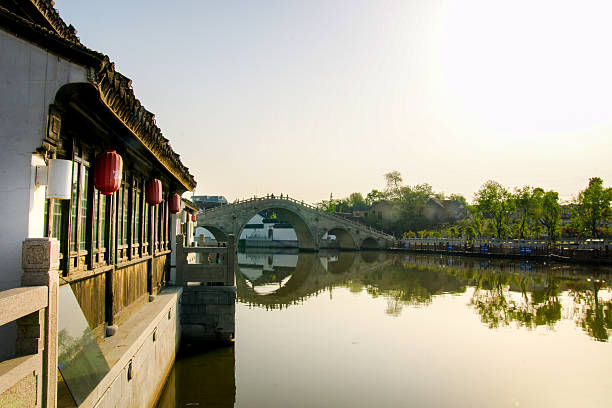 A tranquil scene in Suzhou featuring a traditional stone arch bridge reflected in a calm canal. The setting sun casts a warm glow on the historic architecture and serene waters, typical of Suzhou's classical water towns.