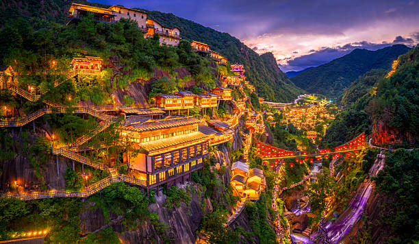 istock Illuminated ancient village with a red lantern bridge and waterfall built into a steep cliff in a green mountain valley at twilight in Jiangxi. 2241411488