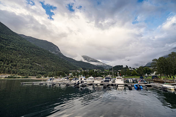 A serene marina rests on a calm fjord in Norway, its docked boats reflected in still water. Steep, forested mountains rise behind the scene, partially shrouded in mist and low clouds. Small buildings nestle among the trees, hinting at a quiet village tucked into the valley. The soft, overcast light enhances the sense of stillness.Shot with Canon R5.
