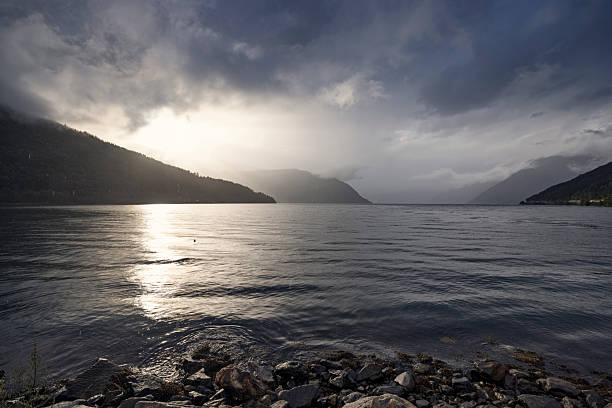 Rain-slicked rocks and tangled seaweed line the edge of a Norwegian fjord, their textures glistening beneath a sky split by sunlight and dense, low-hanging clouds. Gentle ripples move across the water, reflecting shafts of light that pierce the dark overcast. Forested hills fade into mist, adding depth and softness to the scene. The waving water is dotted with drizzle.Shot with Canon R5.