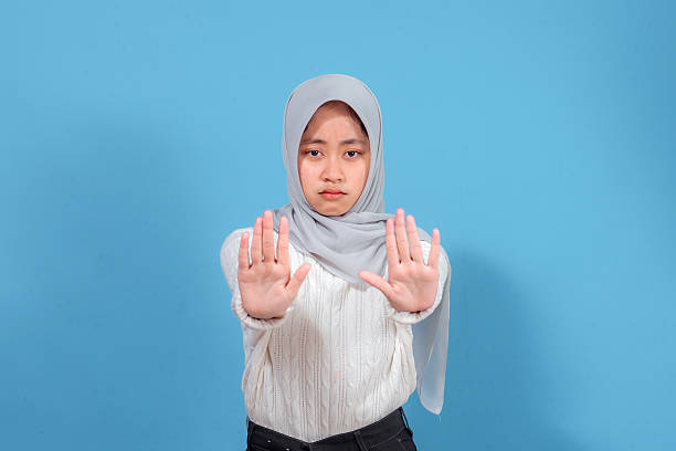 istock Young Asian Muslim woman in a hijab showing a serious expression while raising both hands in a stop gesture, symbolizing refusal, warning, or defense, isolated on blue background. 2241436104