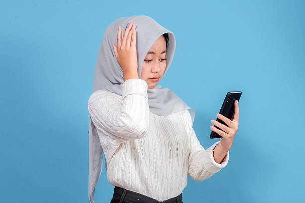 Young Asian Muslim woman wearing hijab and white sweater looking stressed while checking smartphone, touching head in frustration, perfect for communication or technology themes.