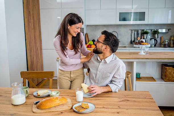 istock Young couple enjoying breakfast in a bright modern kitchen. The man sits at the table while his wife lovingly hugs him from behind. Cozy home atmosphere 2241439606