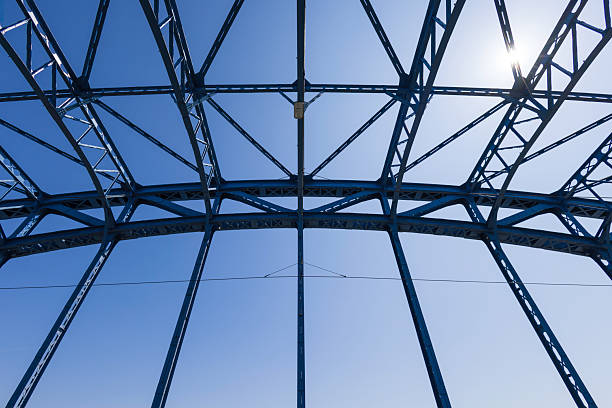 A dramatic low-angle view of a blue steel bridge's lattice framework against a clear sky, highlighting architectural precision, engineering design, and strong geometric patterns.