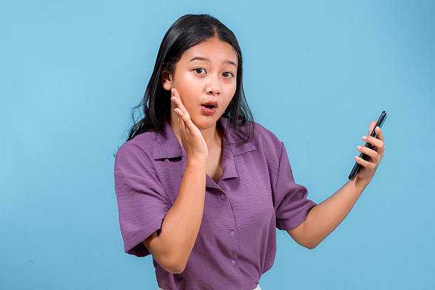 Surprised young Asian woman holding a smartphone, touching her cheek with a shocked expression. Perfect for advertising themes like news, notifications, or unexpected messages.