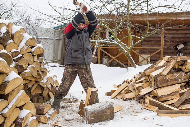 Lumberjack chopping logs in front of an old country house on a winter day. He is wearing yellow protective gloves.