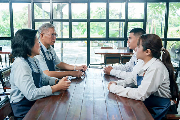 istock Senior asian restaurant owners discussing work strategy with young employees across table teamwork communication collaboration small business planning management partnership leadership meeting 2241455441