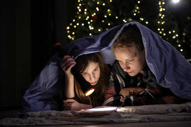 Two girls reading under a blanket with a flashlight near a Christmas tree decorated with glowing lights stock photo