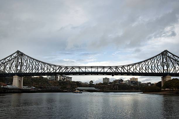 Dramatic suspension bridge spanning a scenic waterway under a cloudy urban skyline.
