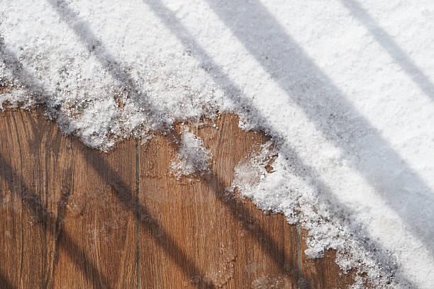Snow blankets a wooden surface, with shadows cast by bars above.