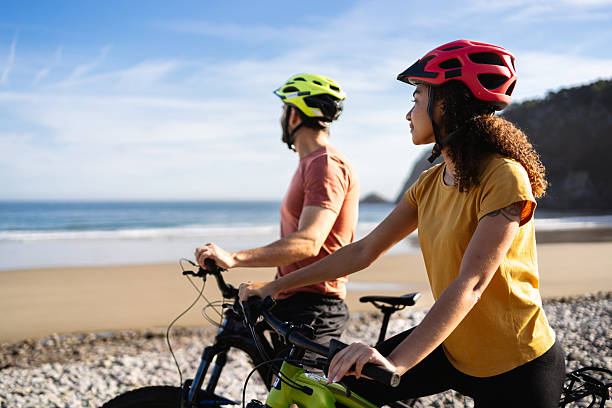 Couple riding electric bikes on a beach, wearing helmets and looking at the ocean. Enjoying a healthy outdoor activity