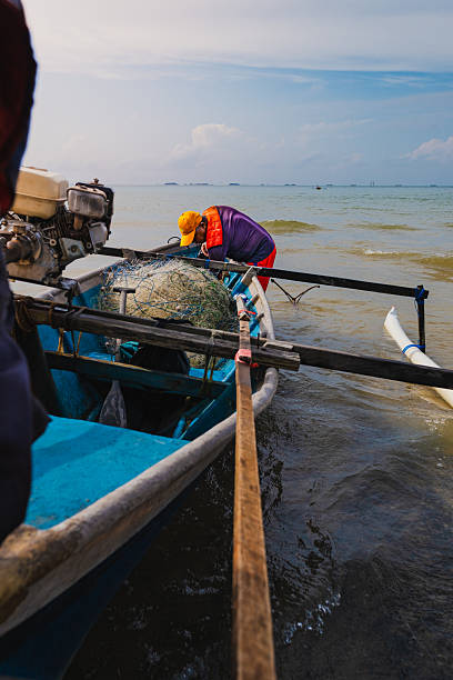 Balikpapan, Indonesia - September 20 2025. Local fisherman arranging tangled fishing nets on a small traditional outrigger boat by the shore under morning sunlight.