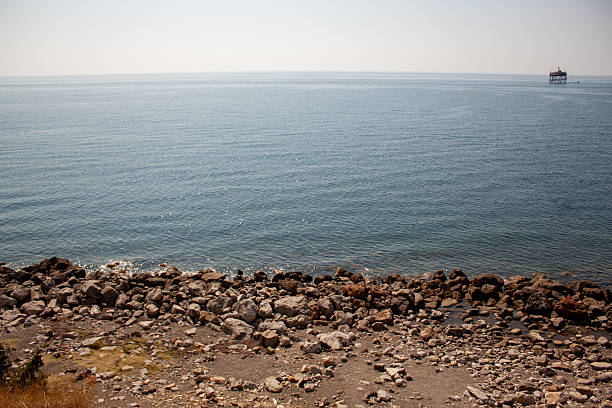 A peaceful coastal scene with rocks along the shoreline. The water is calm and clear with distant views of the horizon under a light blue sky.