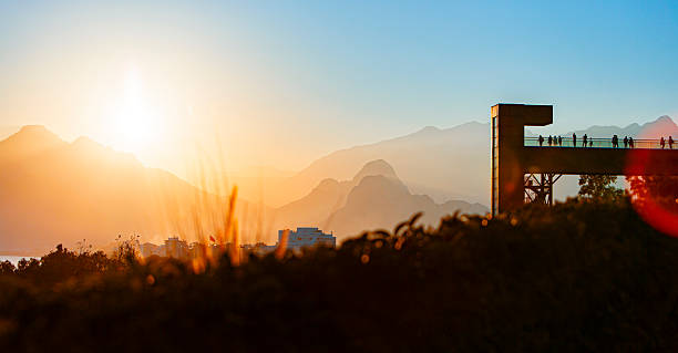 Scenic winter sunset in Antalya, Turkey, with silhouettes of people on a pedestrian bridge overlooking the city. The sun casts a golden glow across the mountains and urban landscape, while trees in the foreground hint at the season’s cooler tones. Ideal for visuals capturing Mediterranean winter charm, tranquil city moments, and the warmth of sunset light during December.