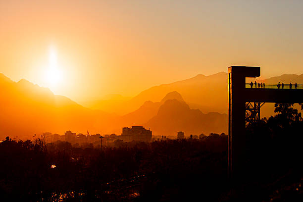 Scenic winter sunset in Antalya, Turkey, with silhouettes of people on a pedestrian bridge overlooking the city. The sun casts a golden glow across the mountains and urban landscape, while trees in the foreground hint at the season’s cooler tones. Ideal for visuals capturing Mediterranean winter charm, tranquil city moments, and the warmth of sunset light during December.