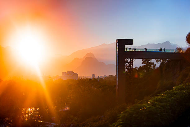 Scenic winter sunset in Antalya, Turkey, with silhouettes of people on a pedestrian bridge overlooking the city. The sun casts a golden glow across the mountains and urban landscape, while trees in the foreground hint at the season’s cooler tones. Ideal for visuals capturing Mediterranean winter charm, tranquil city moments, and the warmth of sunset light during December.