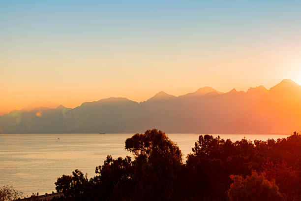 Warm winter sunset over Antalya’s coastline, with rugged cliffs bathed in golden light and a calm sea reflecting the sky’s soft pink and blue tones. Across the water, high-rise buildings line the city’s edge, illuminated by the fading sun. A small boat drifts near the shore, adding motion to the tranquil scene. Ideal for visuals capturing Mediterranean winter beauty, urban-nature contrast, and peaceful coastal moments.