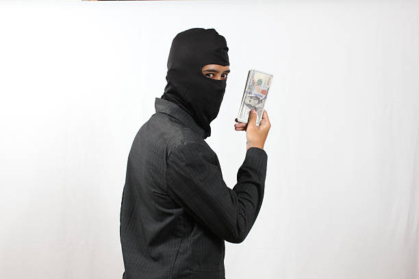 istock A thief wearing a Balaclava mask and a black suit looks back while holding a stack of dollar bills. 2241500523