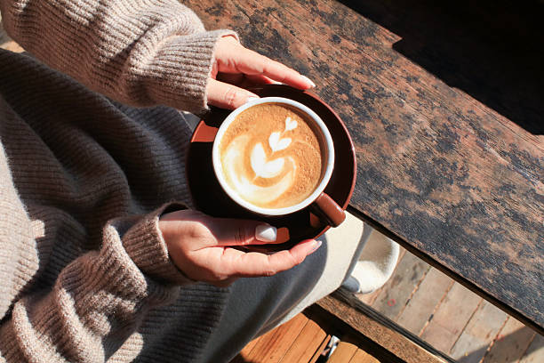 Hand holding hot coffee with latte art on wooden table under sunlight. Autumn Morning breakfast. Creamy coffee cup aromatic.