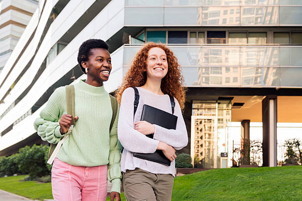 Two happy students, one young man and one young woman, walk in front of a modern glass building on campus, carrying study materials and looking casual and confident.