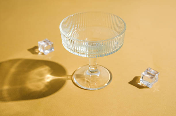 Clear glass coupe with ribbed rim and ice cubes on a muted gold surface with light shadows