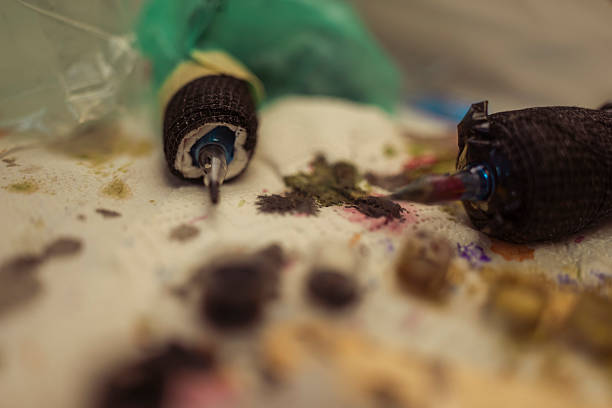 Macro shot of two tattoo machine needles resting on a surface with various colorful inks. The image highlights the tools and pigments used in the tattooing process in a studio setting.