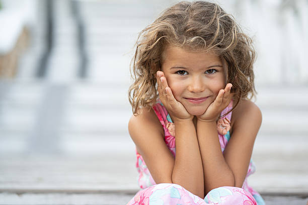 Curly-haired girl resting chin on hands, smiling at camera in summer light — cheerful, carefree elementary-age portrait conveying innocence, daydreaming and joyful youth