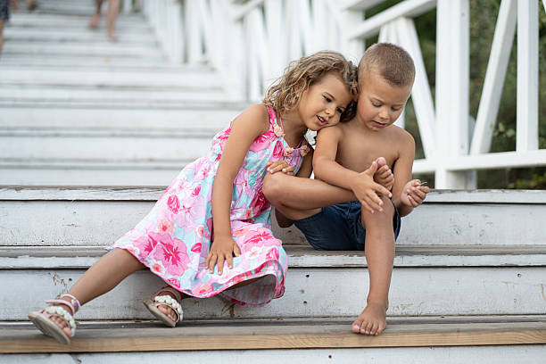 Two children, a girl in a dress and a boy in shorts, spending happy summer moments together, laughing and smiling brightly while sitting on weathered white wooden stairs