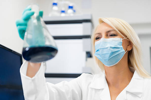 istock Scientist in face mask observing blue solution in flask in lab 2241535388