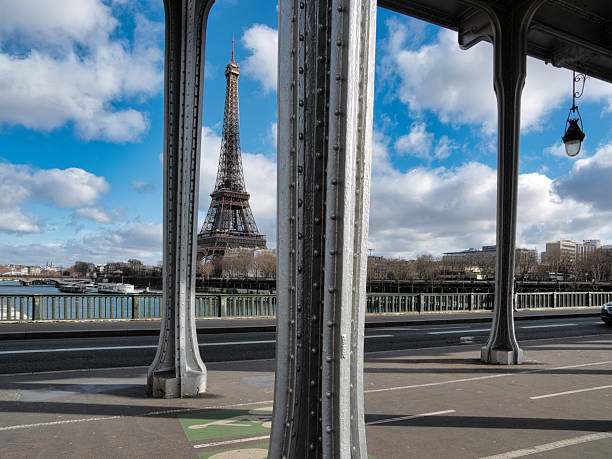 istock Eiffel Tower View from Bir-Hakeim Bridge - The Eiffel Tower prominently featured against a bright blue sky with white clouds, framed by the grey metal structure and lampposts of the Pont de Bir-Hakeim in Paris. 2241547668