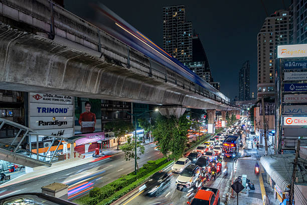 Bangkok, Thailand - 14 October 2025 - City commuters move through traffic under an elevated railway at night, with modern infrastructure, illuminated signs, and motion blur capturing fast-paced life