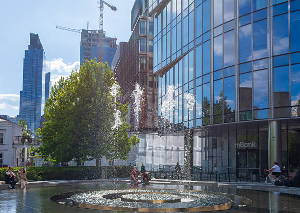 Warsaw, Poland – May 25, 2025: A refreshing city fountain sparkles in sunlight beside glass office towers. People sit nearby, enjoying a warm afternoon surrounded by greenery and modern architecture.