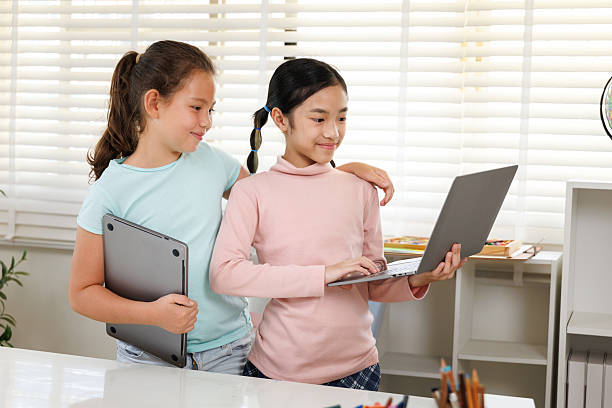 istock Two girls are standing with laptops in a classroom or study room with a large window. One is typing, while the other smiles and holds her computer, 2241562356