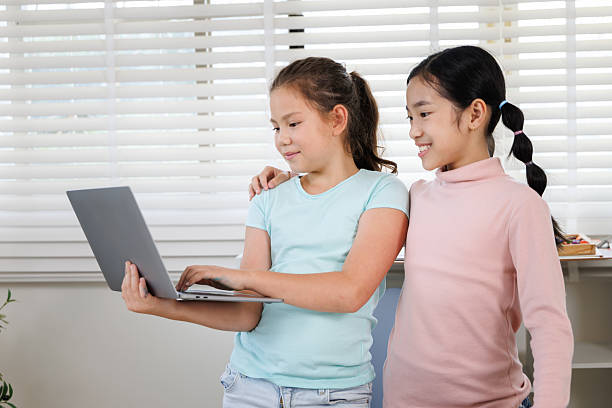 istock Two girls are standing with laptops in a classroom or study room with a large window. One is typing, while the other smiles and holds her computer, 2241562370