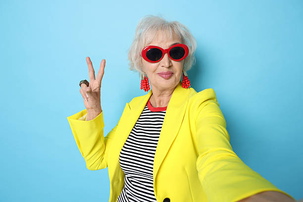 istock Cool grandmother showing V-sign while taking selfie on light blue background 2241563132