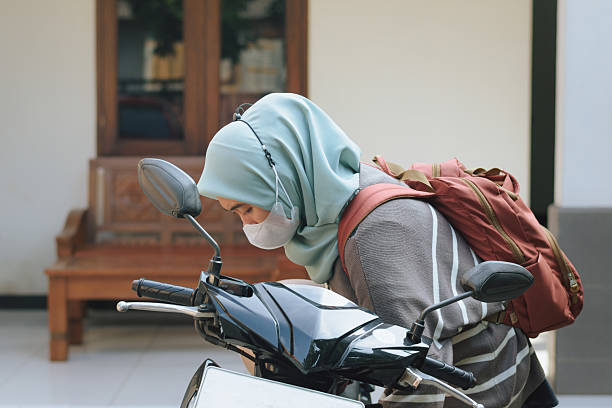 A dedicated young Muslim woman, wearing a hijab and a face mask, performs her morning routine by wiping her motorcycle helmet and carefully fastening the strap before commuting to work