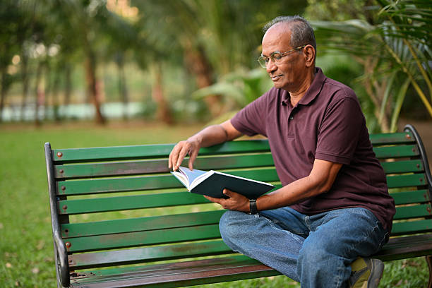 An elderly man sits comfortably on a green park bench, absorbed in a book during golden hour. He reads peacefully amid lush greenery—capturing themes of lifestyle, lifelong learning, mindfulness, and relaxation outdoors.