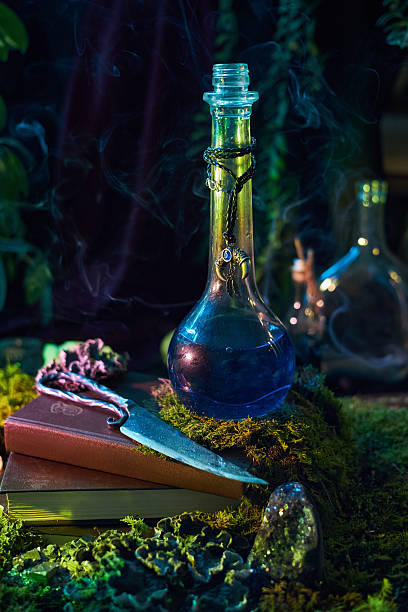 istock glass bottle filled with a blue liquid emits gentle smoke. It sits on a moss-covered surface, surrounded by magical artifacts, stones, and a book, all illuminated by soft green light 2241577878