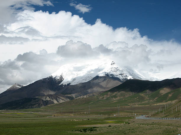 A majestic snow-capped mountain stands prominently with a vast grassland below under a sky filled with clouds.