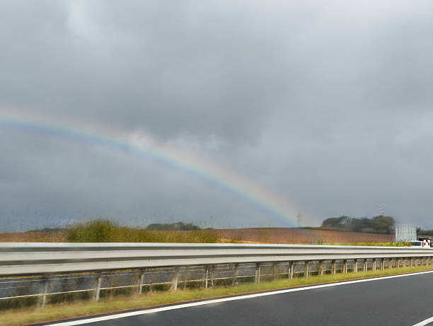 Autumn landscape features a vibrant rainbow over a highway, surrounded by dark clouds and fields, creating a striking visual contrast