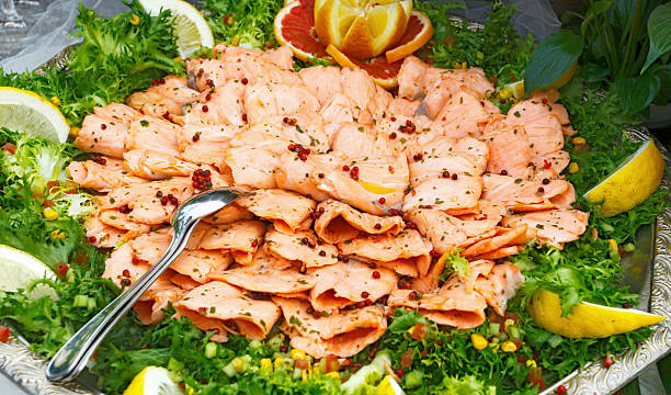 Displaying a platter of delicate salmon fillets garnished with fresh herbs, pink peppercorns, lemon wedges, and grapefruit slices, perfect for a buffet or elegant catering event
