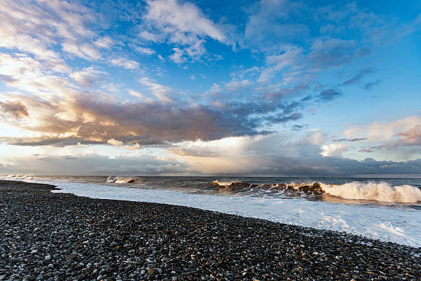 A pebbled shoreline meets rolling waves under a dramatic blue sky with golden sunset light. The scene conveys calm yet energy, ideal for travel, nature, and coastal lifestyle visuals.