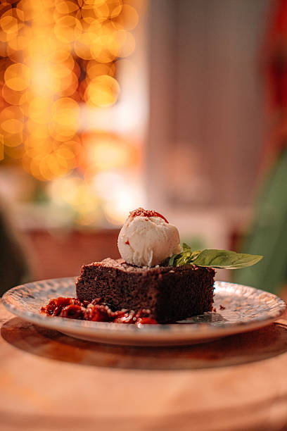 A plate of chocolate brownie topped with a scoop of ice cream, a mint leaf and a cherry against the background of golden garland lights