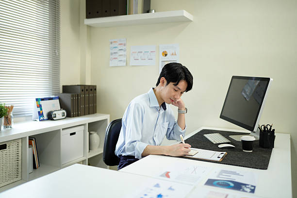 istock Young man reading documents at desk in modern office with computer and bookshelf nearby, showcasing a professional work environment under soft natural light. 2241606174