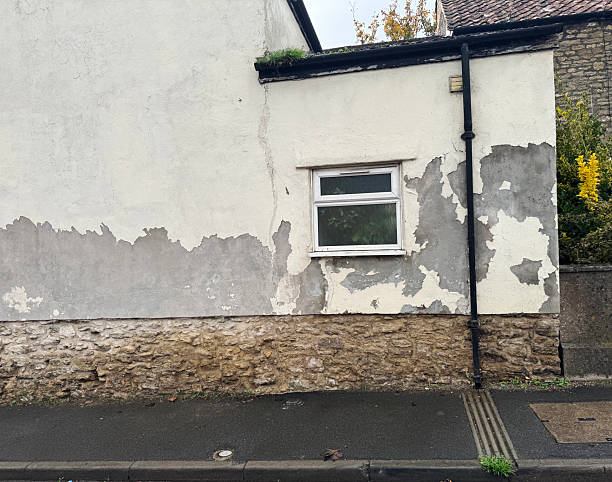 An exterior wall of a building with a white painted surface that is peeling and flaking off, revealing a darker grey layer underneath. The lower portion of the wall is constructed from rough, light-colored stone.