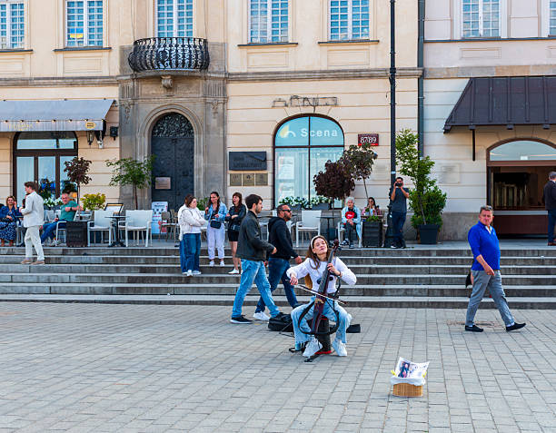Warsaw, Poland – May 25, 2025: A woman performs on the cello in front of a historic building while passersby stop to watch. The performance adds life to the urban atmosphere.