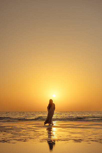 A lone person walks along the wet, reflective sand at sunset, the sky glowing with warm golden hues as the sun nears the horizon. The calm sea and soft light create a peaceful, contemplative mood perfect for travel, lifestyle, and relaxation themes.