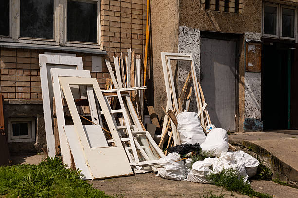 Discarded doors and construction materials are piled near a rundown building. Overgrown grass and debris hint at the neglect of this urban location during the day.